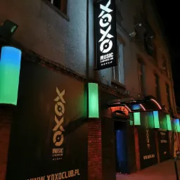 Zewnętrzny widok nocny klubu muzycznego z podświetlanymi kasetonami reklamowymi z logo i nazwą, oraz cylinderami świetlnymi w kolorach niebieskim i zielonym, oświetlającymi wejście.