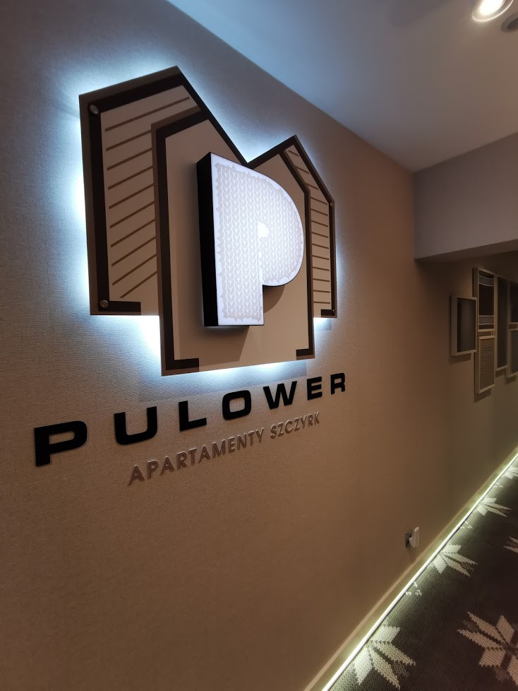 Podświetlany, przestrzenny logotyp 'PULOWER Apartamenty Szczyrk' z motywem dzianiny na literze P, umieszczony na ścianie w korytarzu.