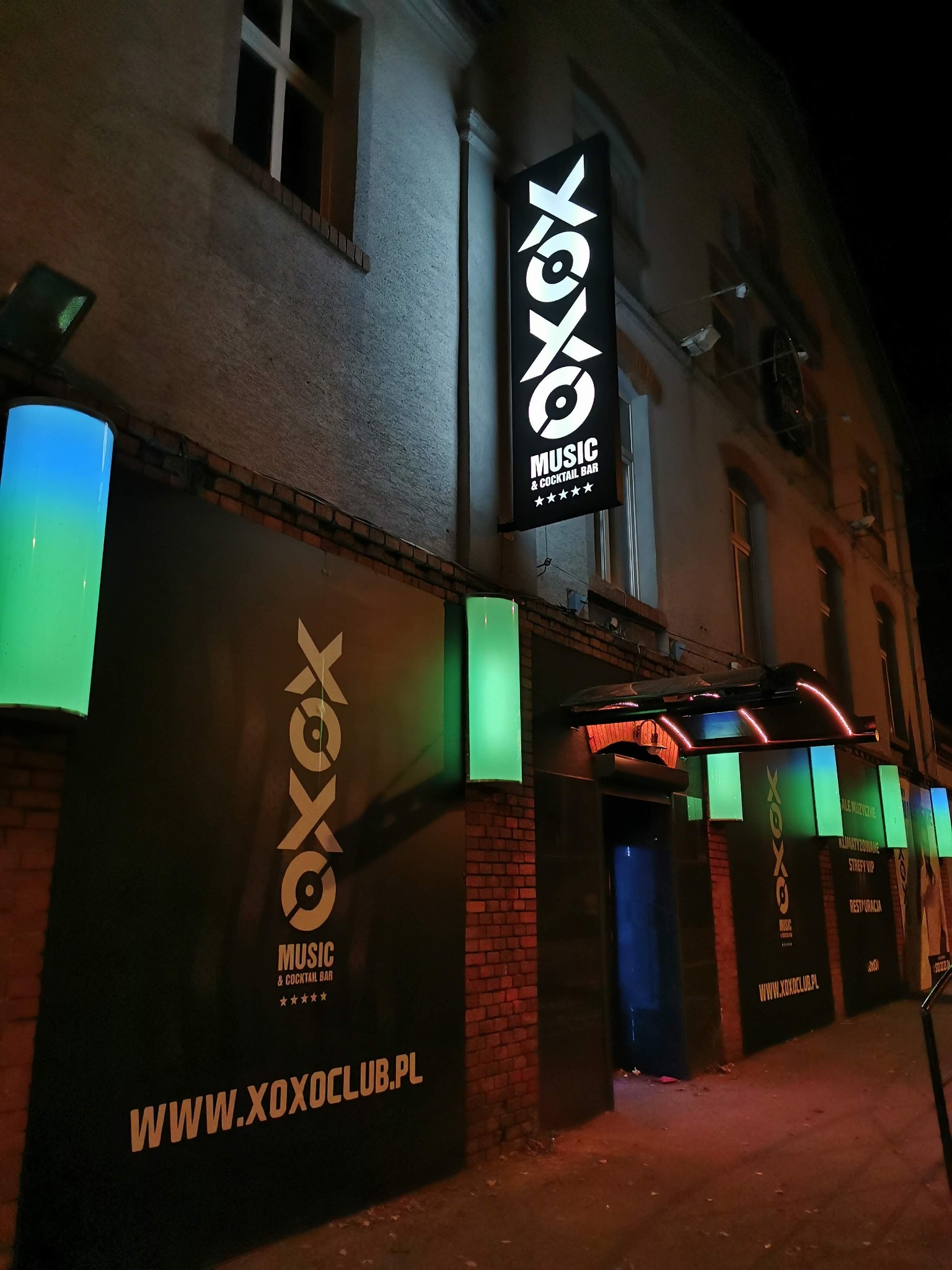 Zewnętrzny widok nocny klubu muzycznego z podświetlanymi kasetonami reklamowymi z logo i nazwą, oraz cylinderami świetlnymi w kolorach niebieskim i zielonym, oświetlającymi wejście.