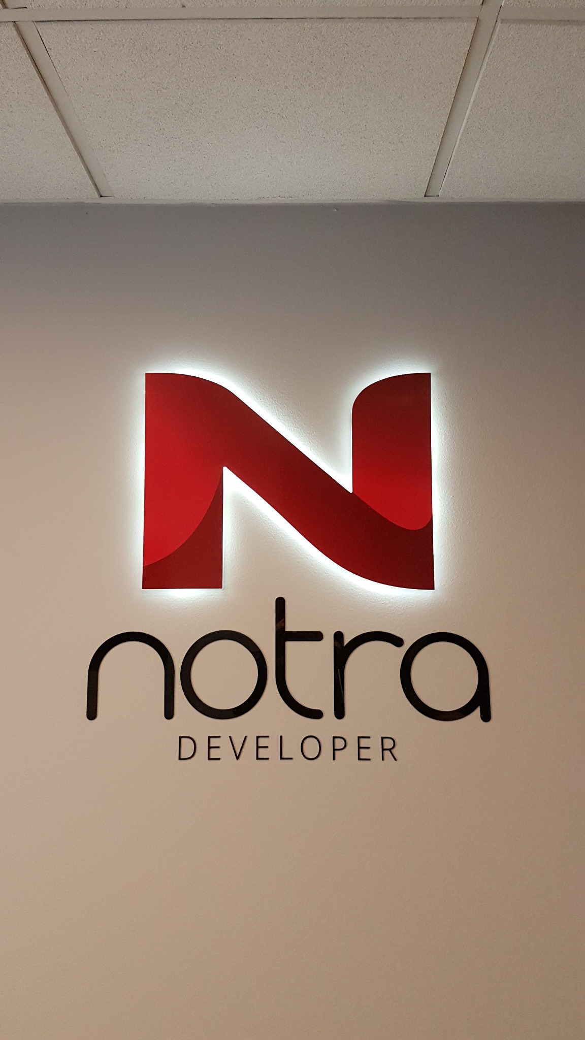 Podświetlane logo 'Notra Developer' z czerwoną literą 'N' na białej ścianie, widoczne elementy sufitu kasetonowego.