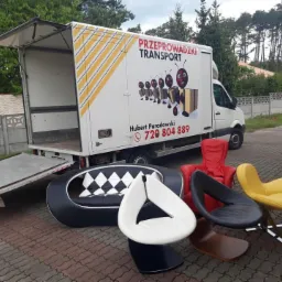 Otwarty samochód dostawczy z rampą, meble (sofa, krzesła) na zewnątrz, logo firmy przeprowadzkowej z mrówkami niosącymi kartony na boku pojazdu.