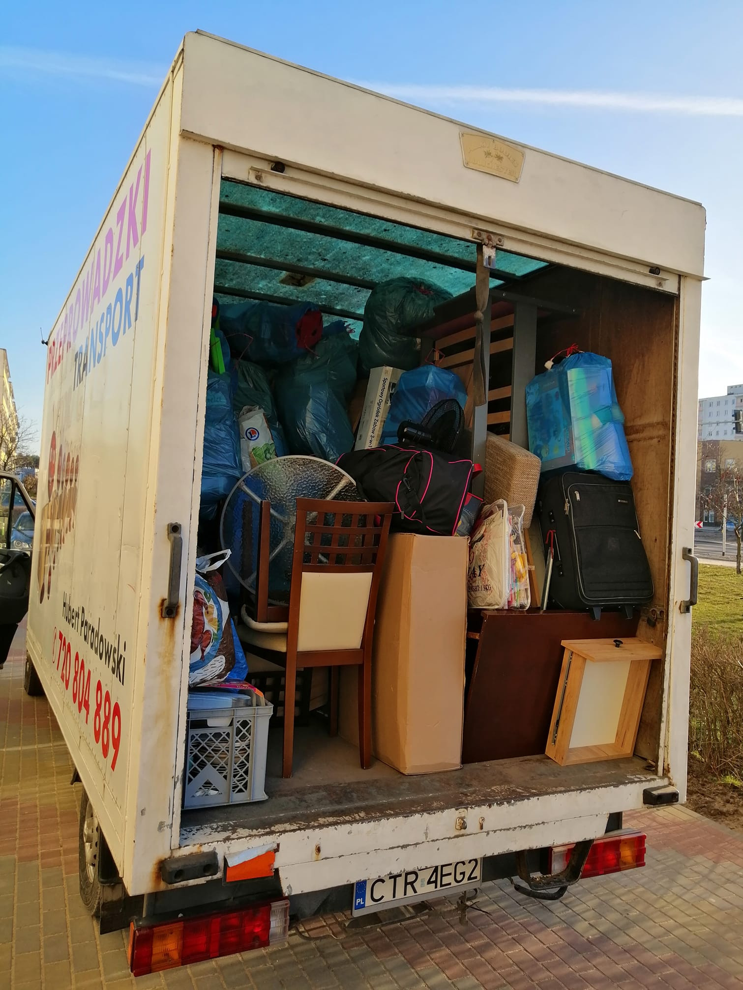 Otwarta przestrzeń ładunkowa białego samochodu dostawczego wypełniona po brzegi różnymi przedmiotami, w tym meblami, torbami i kartonami, przygotowanymi do transportu.