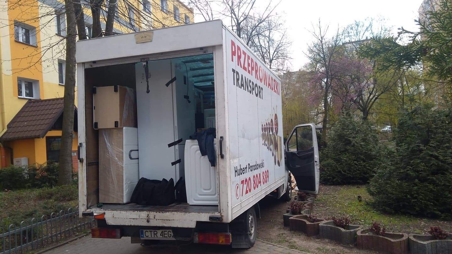 Otwarty tył białej furgonetki z napisem 'PRZEPROWADZKI TRANSPORT' załadowanej kartonami, pralką i lodówką na tle budynku mieszkalnego i zieleni.