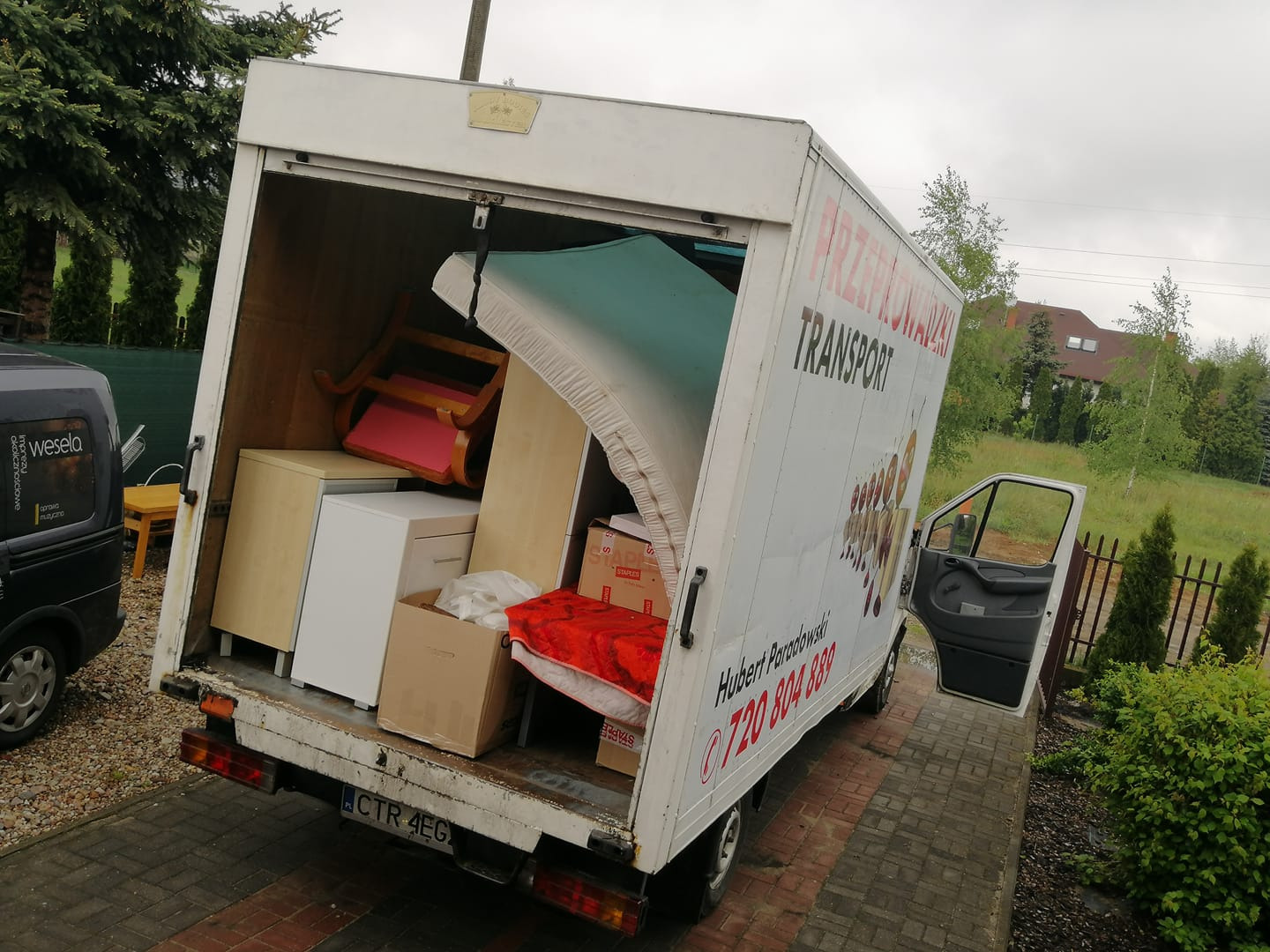 Otwarta paka białego samochodu dostawczego wypełniona meblami i kartonami, widoczny narożnik materaca, krzesło z różowym siedziskiem i biała szafka, na boku pojazdu napis 'PRZEPROWADZKI TRANSPORT'...