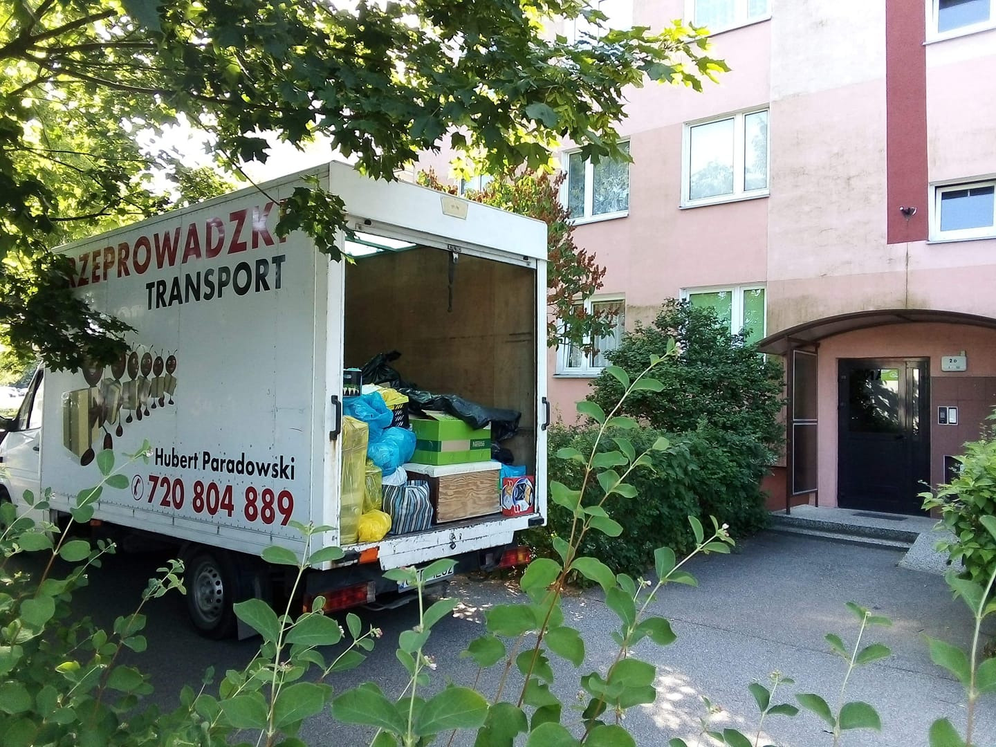 Otwarty samochód dostawczy z napisem 'PRZEPROWADZKI TRANSPORT' i numerem telefonu, załadowany workami i kartonami, zaparkowany przed budynkiem mieszkalnym z zielenią.