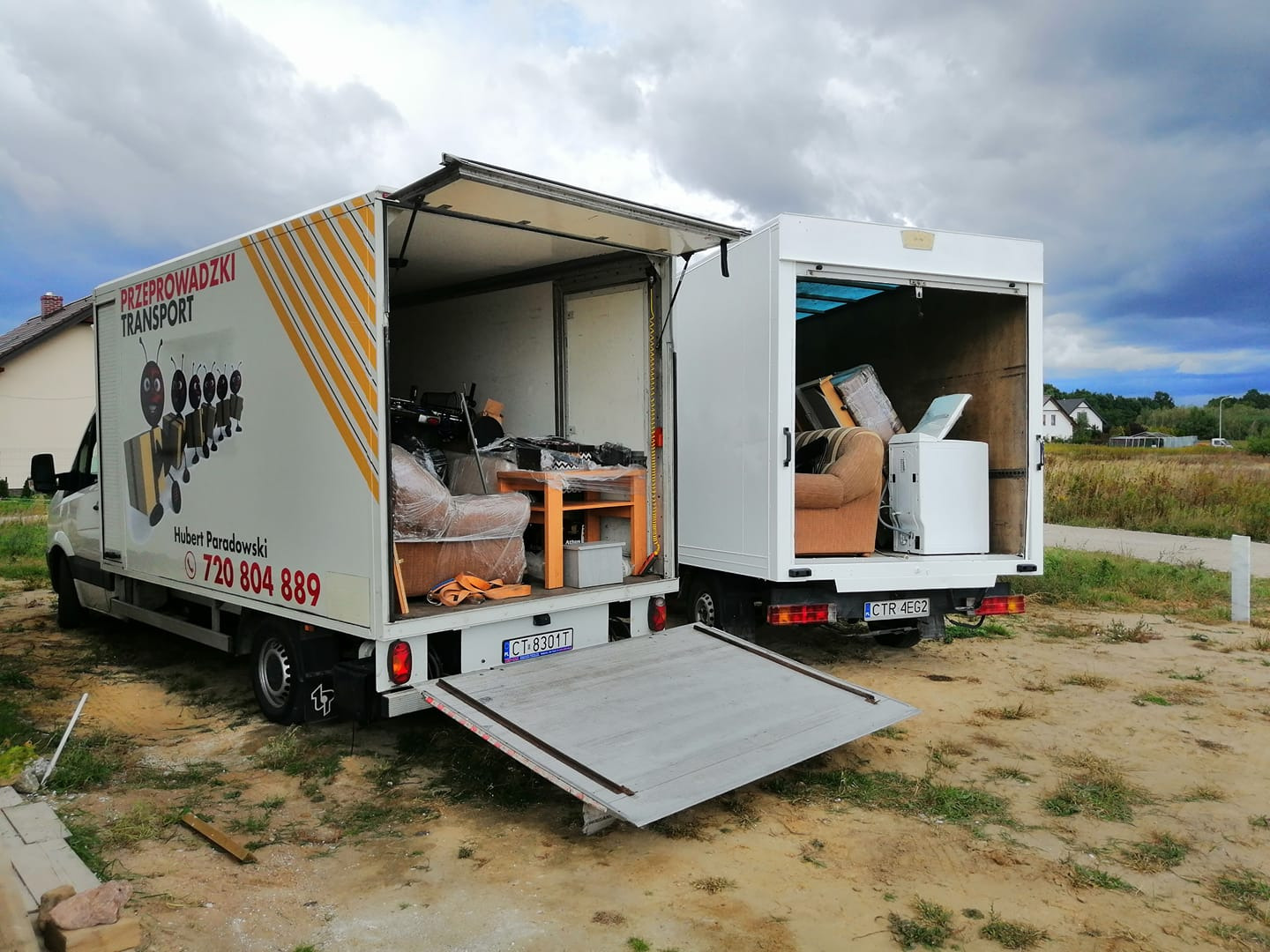 Dwa busy transportowe z otwartymi przestrzeniami ładunkowymi, jeden z reklamą firmy przeprowadzkowej z numerem telefonu, drugi z widoczną pralką i fotelem w środku, sugerujące załadunek...