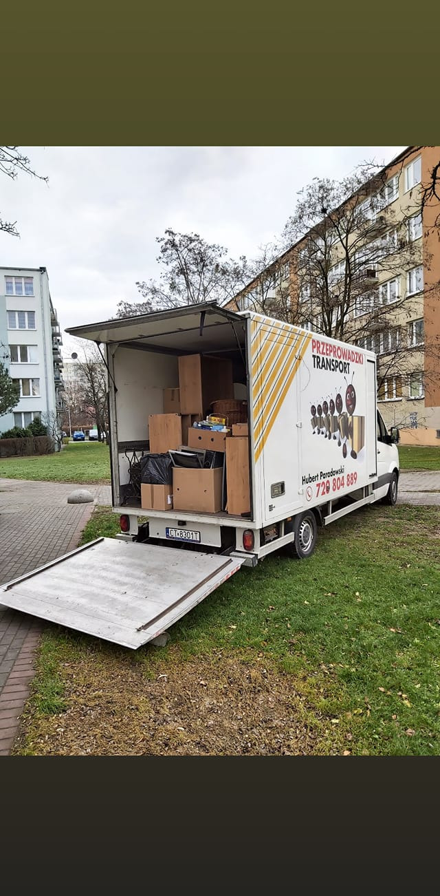 Otwarty samochód dostawczy z kartonami i przedmiotami w środku, zaparkowany na trawniku przed blokiem mieszkalnym; widoczna rampa załadunkowa i logo firmy z numerem telefonu.