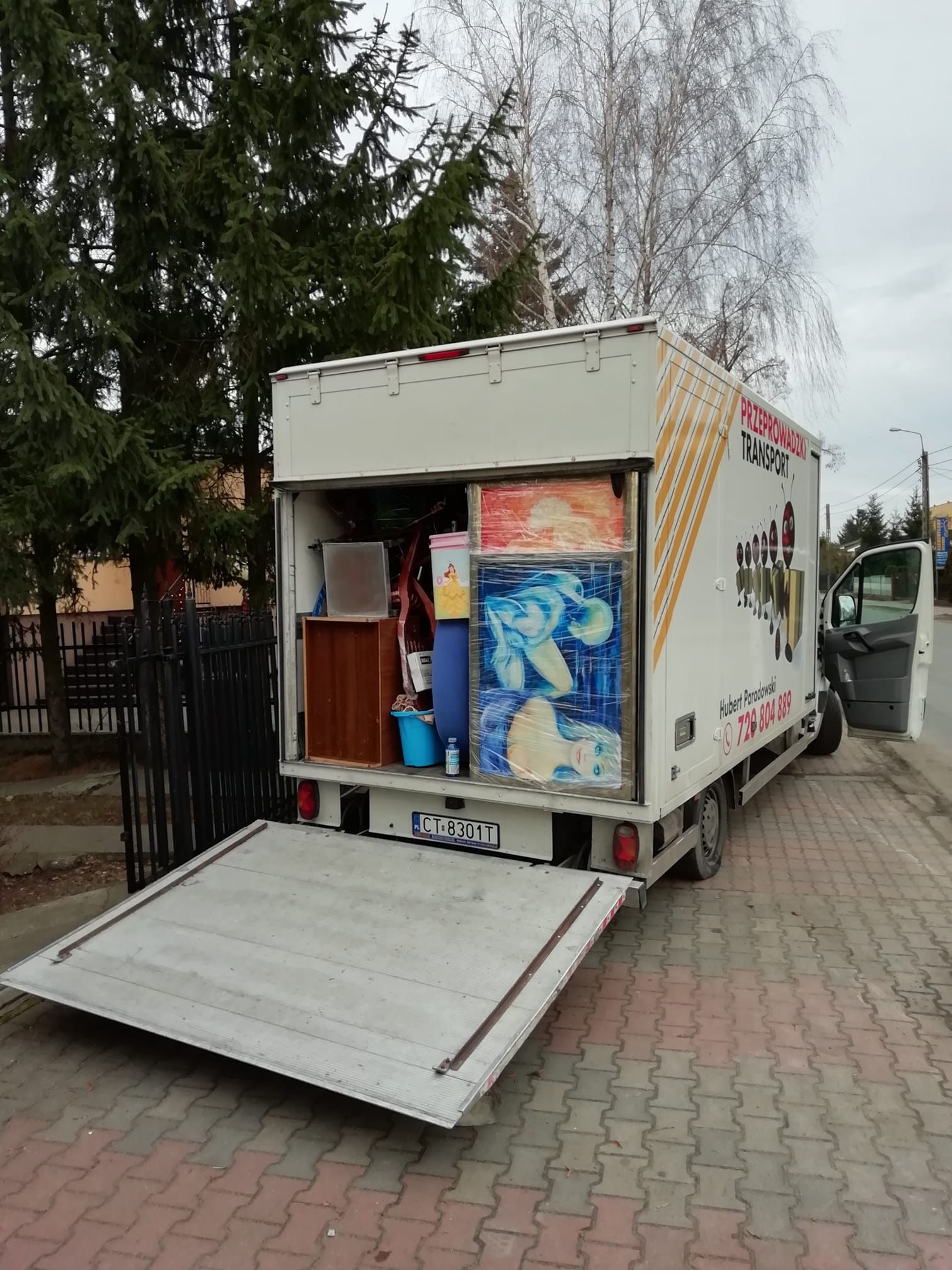 Ciężarówka transportowa z otwartą rampą załadowaną meblami i obrazami, zaparkowana na chodniku.