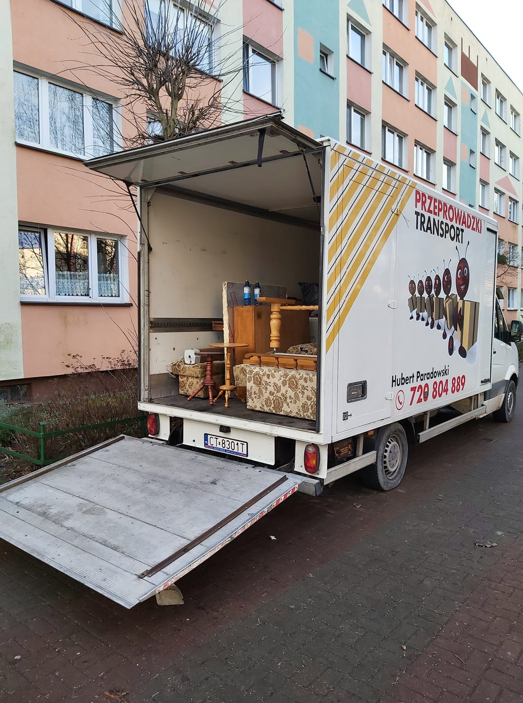 Otwarty furgon z meblami (stół, krzesła, tapczan) podczas załadunku na tle bloku mieszkalnego. Na boku furgonu logo firmy transportowej z numerem telefonu.