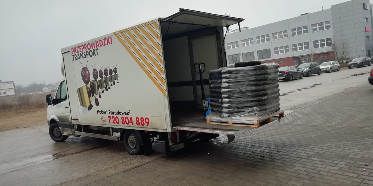 Otwarty tył białego samochodu dostawczego z grafiką mrówek niosących paczki, załadowanego paletą z czarnymi workami, obok niebieski wózek paletowy.