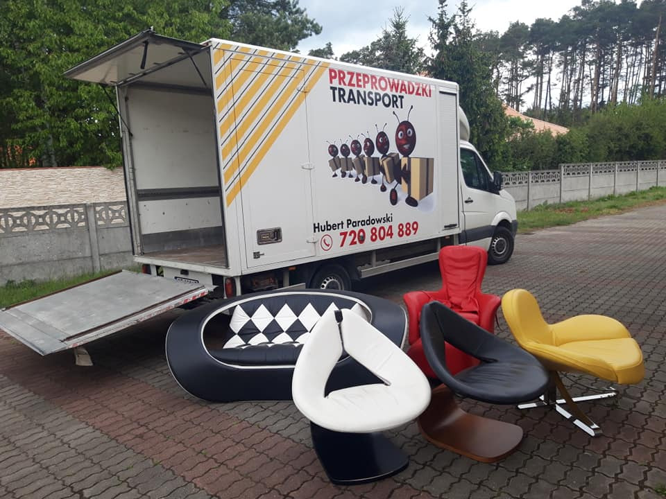 Otwarty samochód dostawczy z rampą, meble (sofa, krzesła) na zewnątrz, logo firmy przeprowadzkowej z mrówkami niosącymi kartony na boku pojazdu.