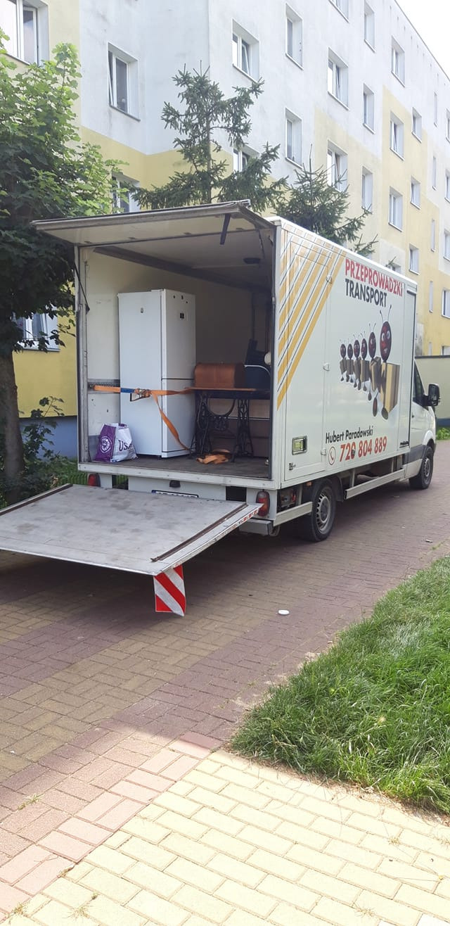 Otwarty samochód dostawczy z wyposażeniem: biała lodówka, stara maszyna do szycia i torba z napisem US, zabezpieczone pasami, na tle bloku mieszkalnego.