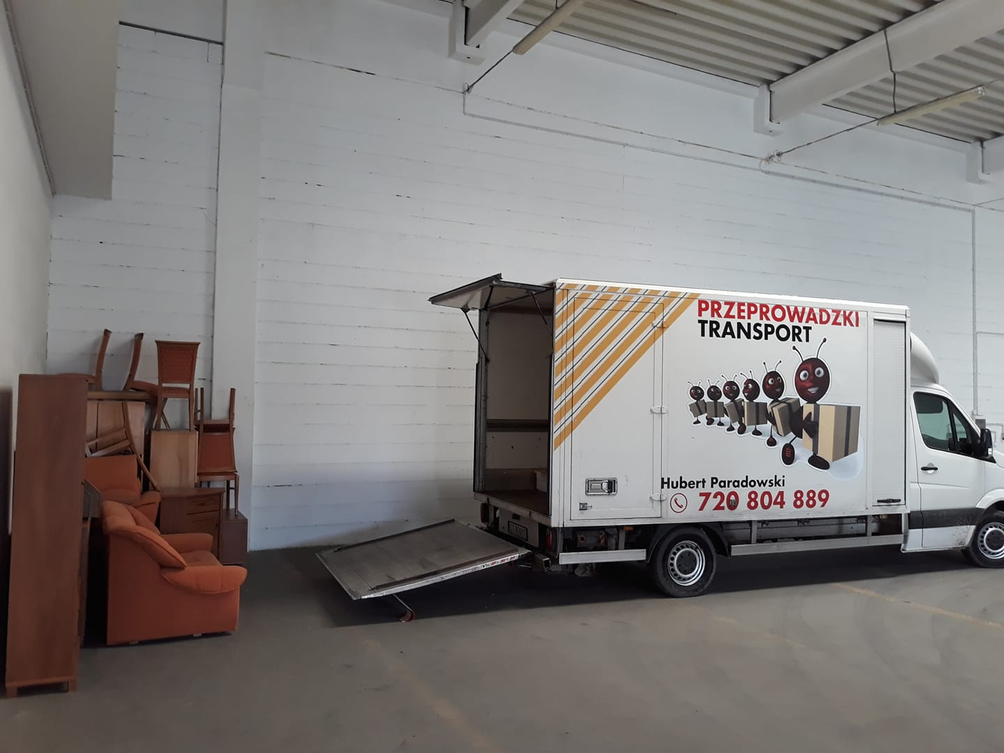 Częściowo załadowany meblami samochód dostawczy z otwartą rampą i logo firmy transportowej z mrówkami niosącymi paczki, zaparkowany w magazynie.