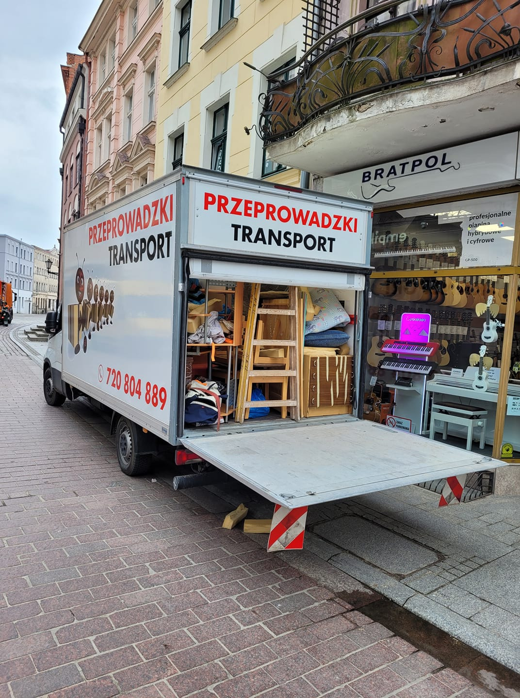 Otwarty samochód dostawczy z napisem 'Przeprowadzki Transport' zaparkowany na ulicy, załadowany meblami i sprzętami, obok sklepu muzycznego Bratpol.