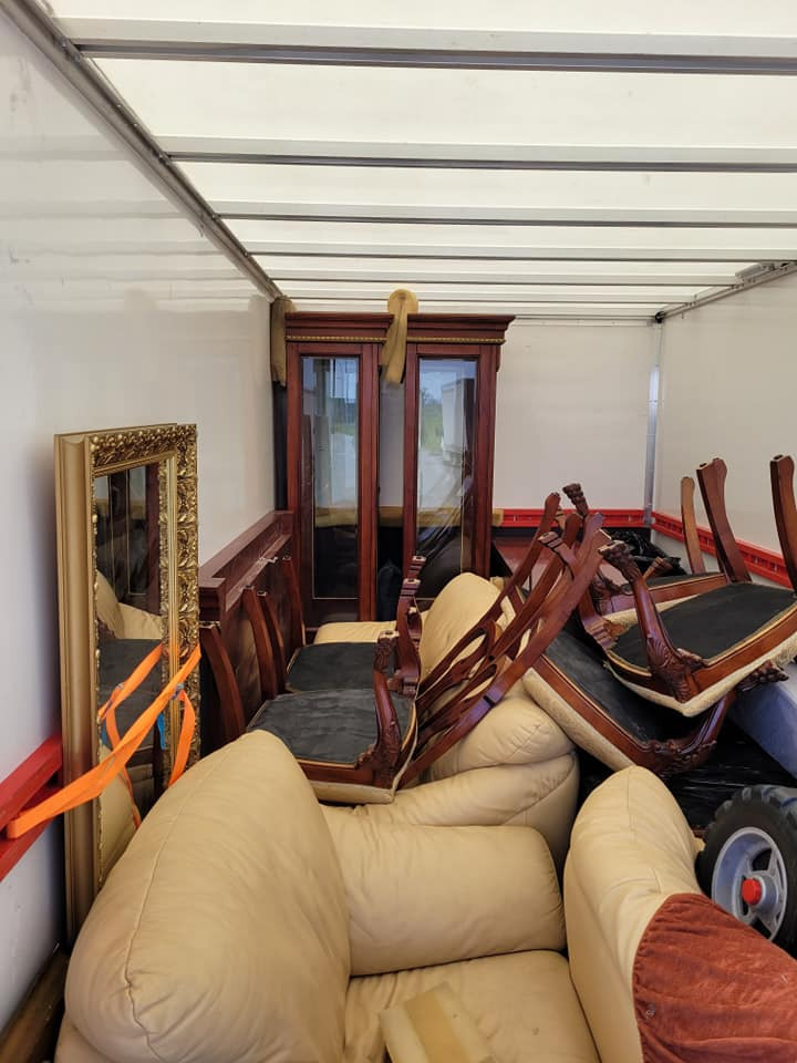 Wnętrze furgonetki załadowane antycznymi meblami: lustro w złotej ramie zabezpieczone pomarańczowym pasem, szafa z ciemnego drewna, krzesła z rzeźbionymi oparciami i beżowa skórzana sofa.