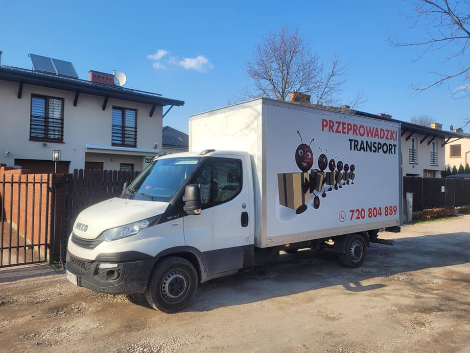 Biała furgonetka z grafiką mrówek niosących paczki i napisem 'Przeprowadzki Transport' zaparkowana na żwirowej drodze przed domem jednorodzinnym w słoneczny dzień.