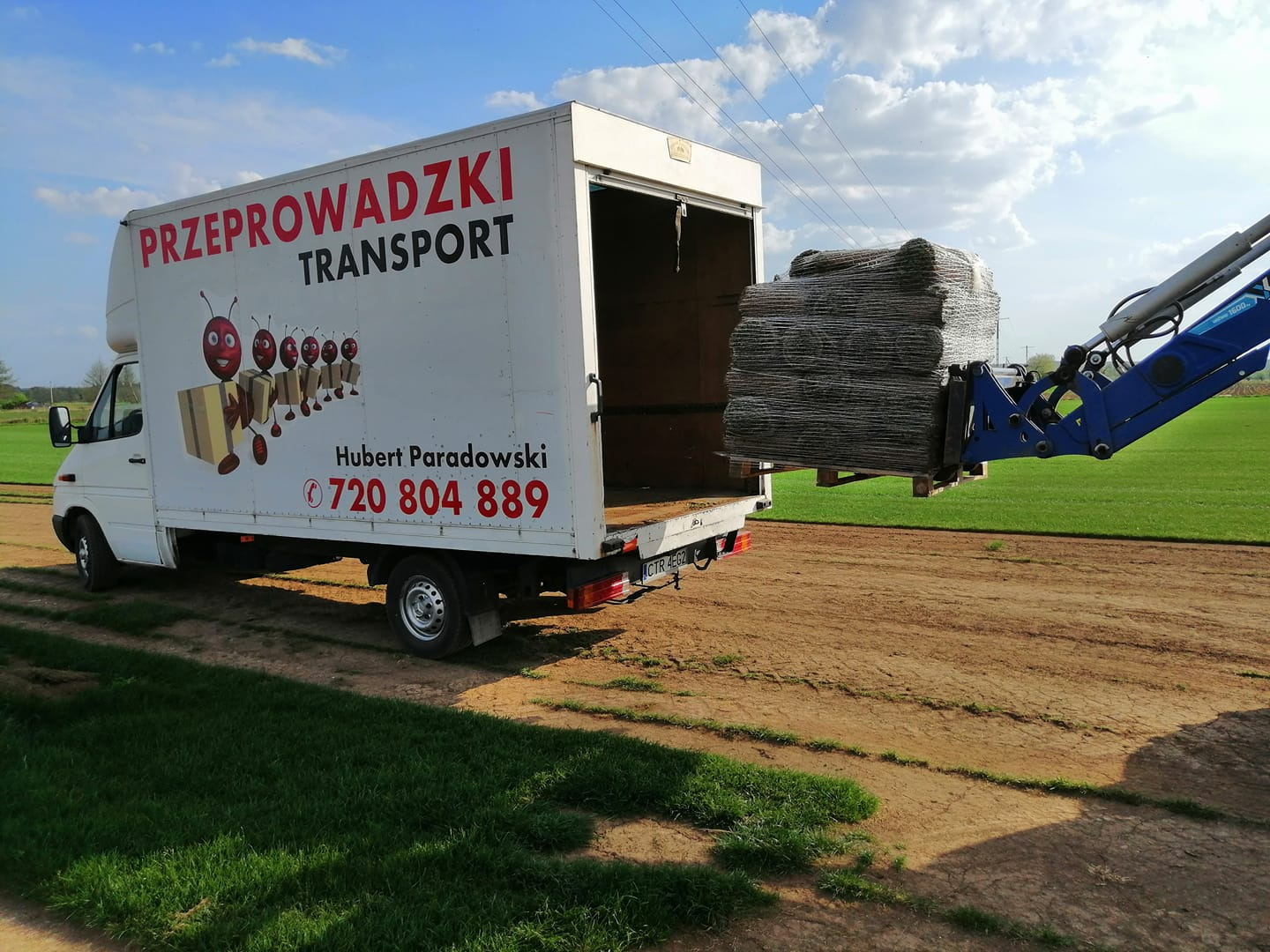 Biała furgonetka z napisem 'PRZEPROWADZKI TRANSPORT' i rysunkiem mrówek niosących kartony, załadowywana za pomocą niebieskiego wózka widłowego paletą z siatką ogrodzeniową na tle zielonego pola...