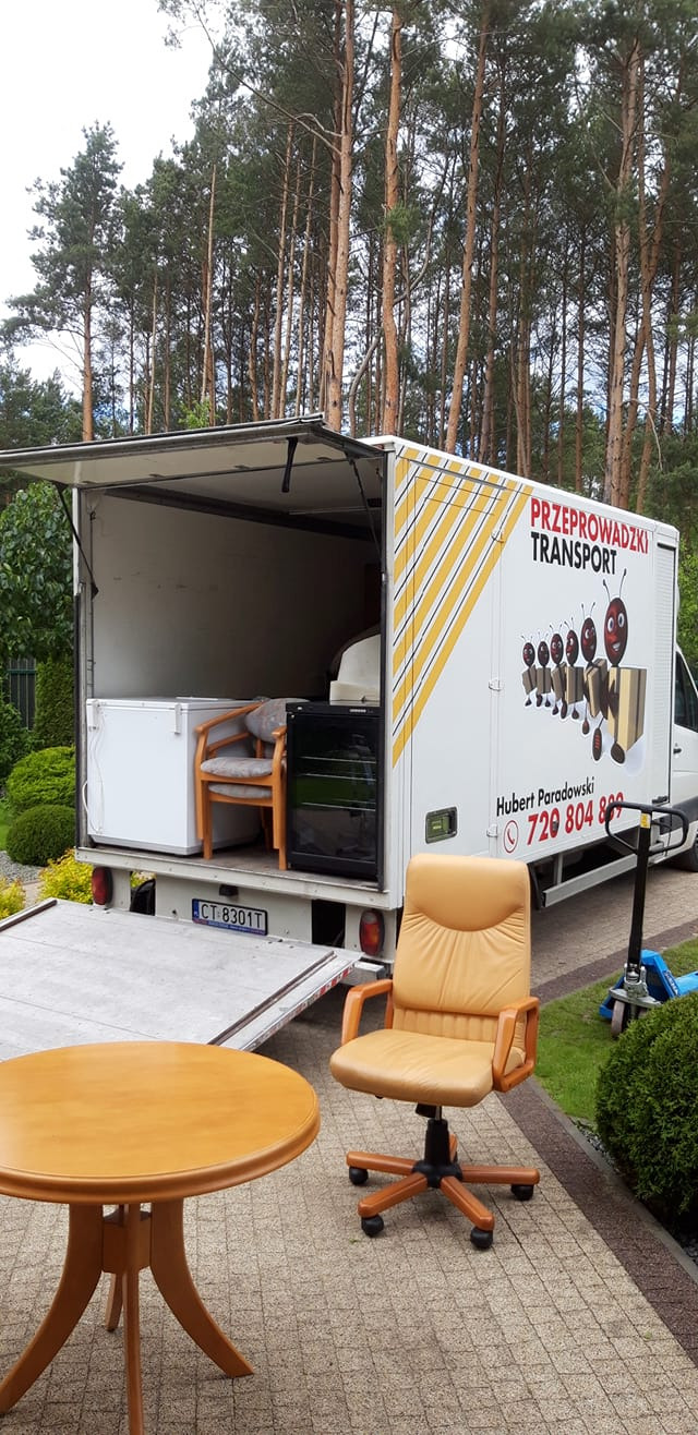 Otwarty furgon z meblami i sprzętem AGD, w tle las, na pierwszym planie okrągły stół i fotel biurowy na kostce brukowej.