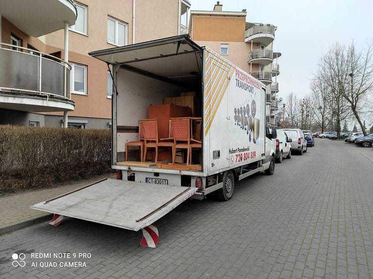 Otwarta paka samochodu dostawczego z meblami (krzesła i szafka) na podnośniku hydraulicznym, zaparkowanego na osiedlu mieszkaniowym; widoczny numer telefonu firmy na boku pojazdu.
