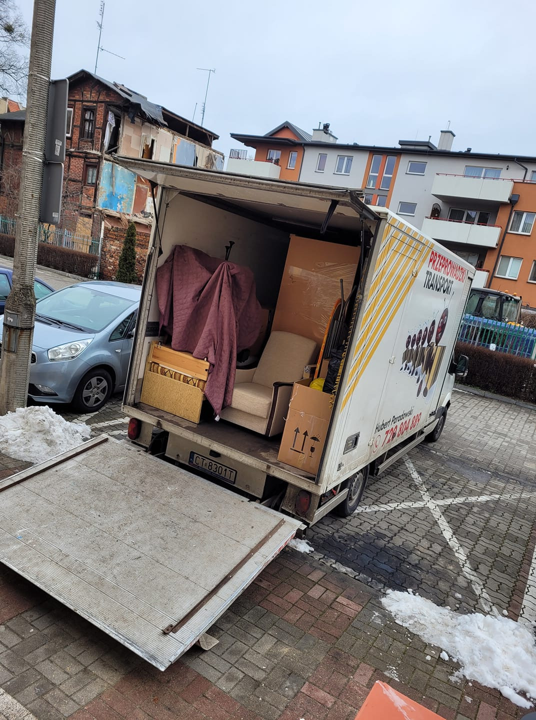 Otwarta paka busa transportowego z meblami (fotel, szafka) zabezpieczonymi folią i kocem, na parkingu z kostki brukowej, w tle budynki mieszkalne i resztki śniegu.