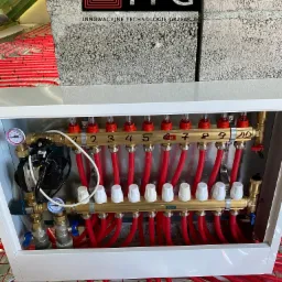 Rozdzielacz ogrzewania podłogowego z czerwonymi rurami, widoczne manometry, numery obieg&oacute;w od 1 do 10, logo ITG na tle bloczk&oacute;w betonowych.