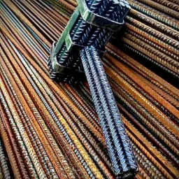 Zestawienie prętów żebrowanych złączonych metalowymi obejmami, ułożonych na tle zardzewiałych prętów.