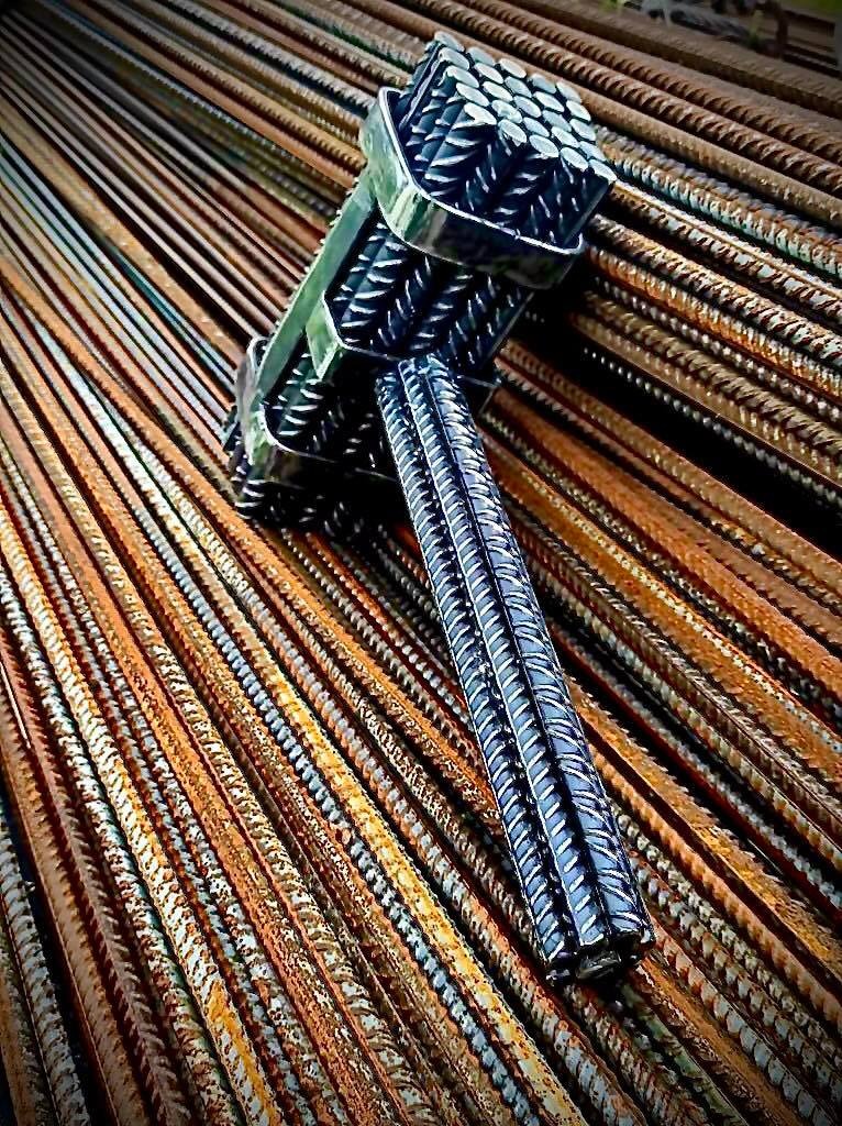 Zestawienie prętów żebrowanych złączonych metalowymi obejmami, ułożonych na tle zardzewiałych prętów.