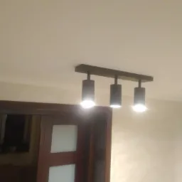 Czarna listwa sufitowa z trzema cylindrycznymi reflektorami LED, oświetlającymi sufit i fragment ściany z widocznym gniazdkiem elektrycznym oraz górną część drewnianych drzwi z białymi panelami.