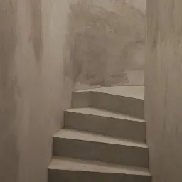 Betonowe schody w surowym wnętrzu z szarymi ścianami o nieregularnej fakturze i punktowym oświetleniem na górze.