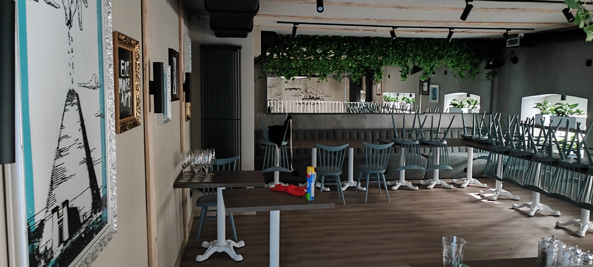 Pusta sala restauracyjna z odwróconymi krzesłami na stołach, przygotowana do sprzątania, widoczne elementy dekoracyjne na ścianach i wiszące rośliny.