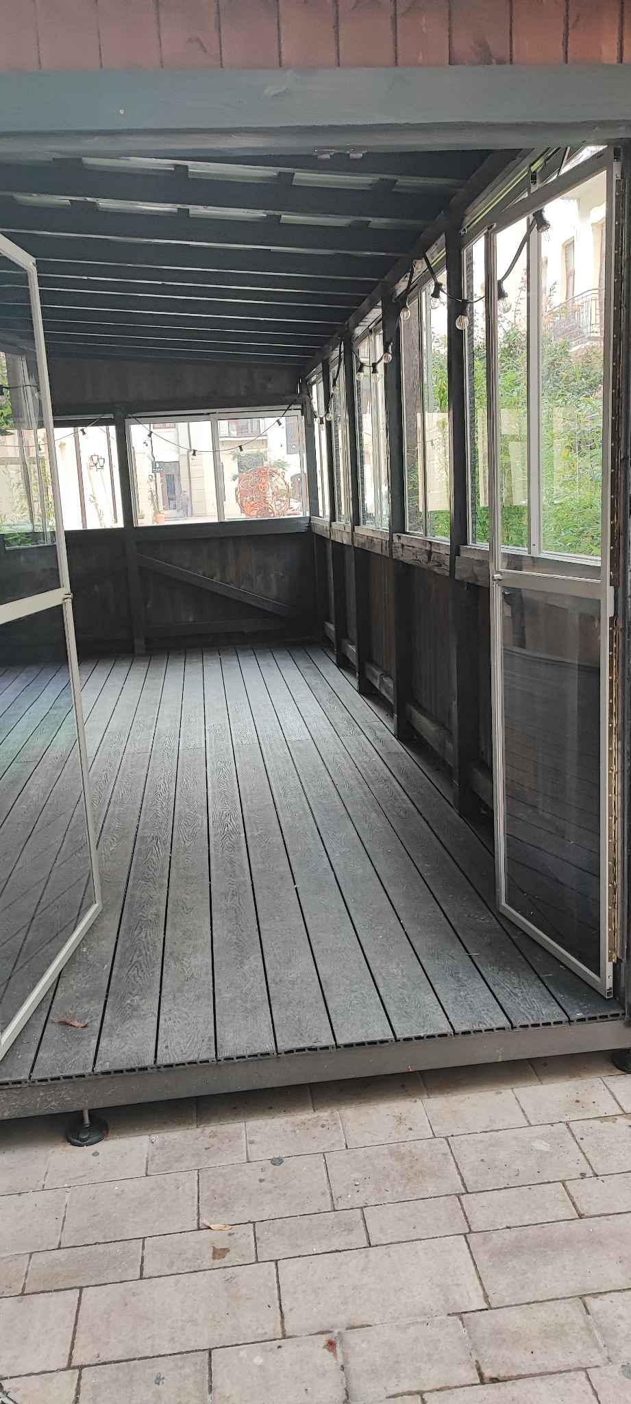 Zadaszony taras z szarą drewnianą podłogą, otwartymi szklanymi drzwiami i widokiem na ulicę z dekoracyjną rzeźbą.