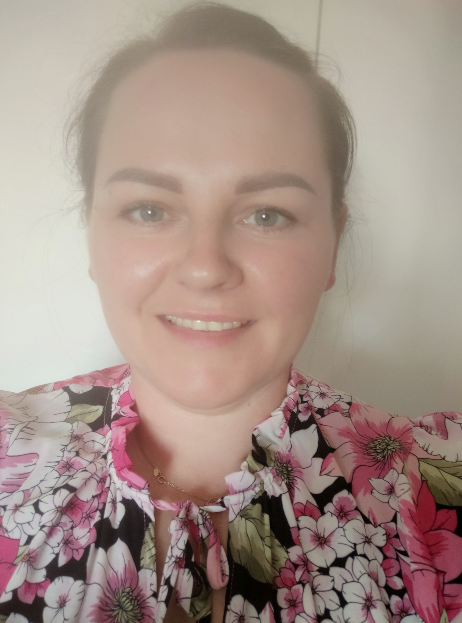 Portret uśmiechniętej kobiety w bluzce w kwiaty, prawdopodobnie opiekunki do dziecka, z delikatnym złotym łańcuszkiem na szyi, na tle białej ściany.