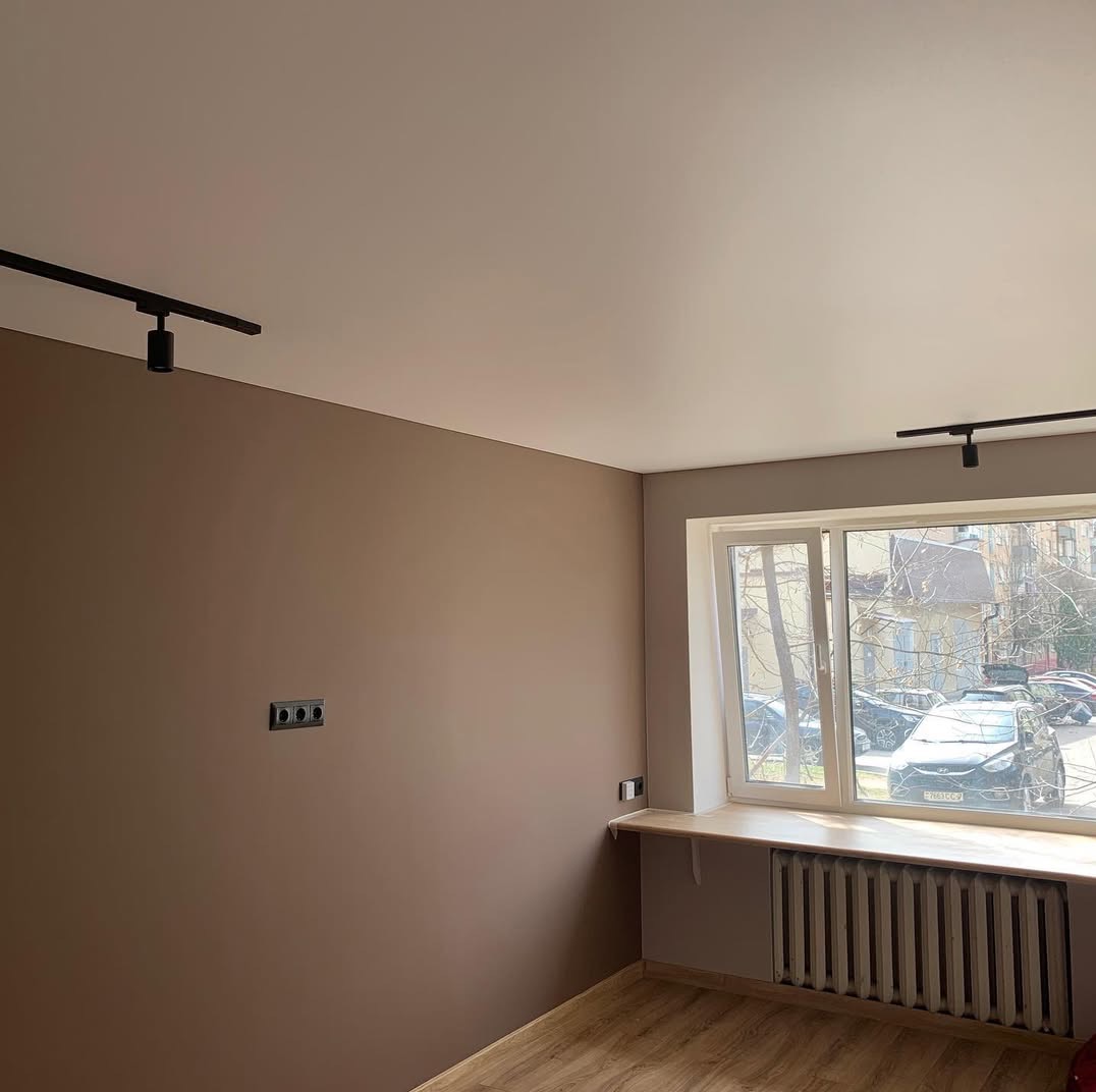 Pomieszczenie po malowaniu ścian na brązowo, z drewnianą podłogą i grzejnikiem pod oknem. Widoczne czarne reflektory sufitowe i gniazdka elektryczne.