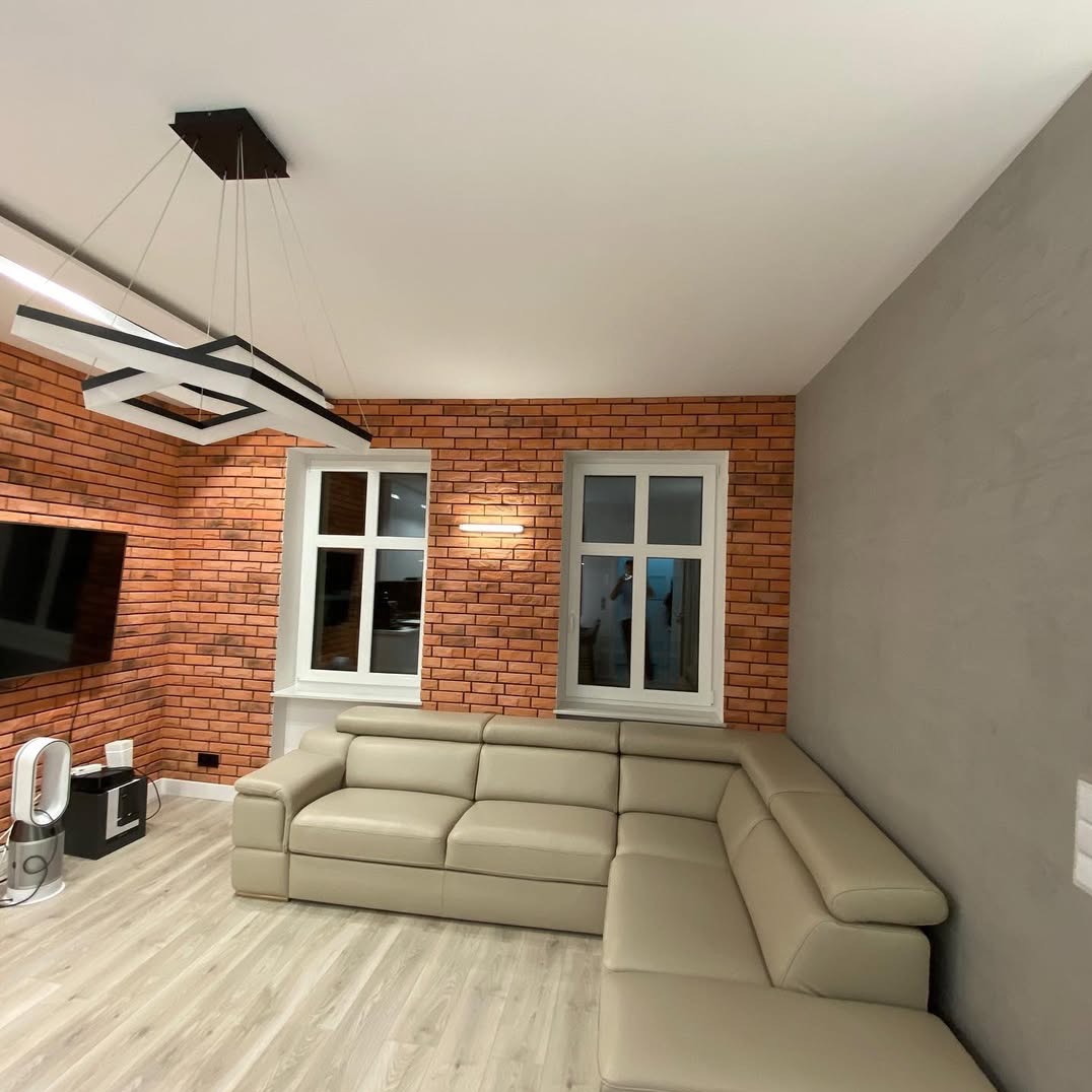 Salon z ceglaną ścianą, białymi oknami i narożną sofą w odcieniach beżu. Nowoczesna lampa sufitowa i jasna podłoga. Ściana pomalowana na szaro.