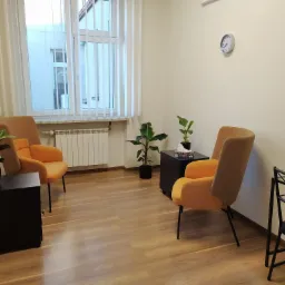 Jasny gabinet z dwoma pomarańczowymi fotelami, małymi czarnymi stolikami, roślinami doniczkowymi, zegarem na ścianie i drewnianą podłogą.