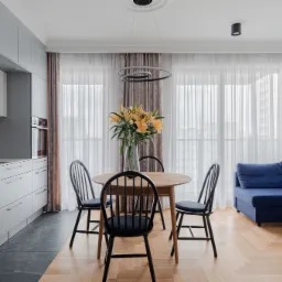 Jasne wnętrze mieszkania z aneksem kuchennym, okrągłym stołem z krzesłami i niebieską sofą narożną, parkiet ułożony w jodełkę, bukiet żółtych lilii w wazonie na stole.