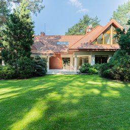 Michał Ziędalski - Rozległy, jednopiętrowy dom z czerwonym dachem, otoczony bujną zielenią drzew i trawnikiem, sfotografowany w słoneczny dzień z wyraźnie widocznymi oknami i kolumnami podtrzymującymi zadaszenie.