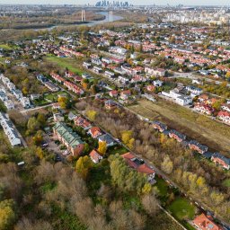 Michał Ziędalski - Panoramiczny widok z lotu ptaka na osiedle domów jednorodzinnych z czerwonymi dachami, w tle rzeka z mostem i panorama miasta z wieżowcami, jesienna kolorystyka drzew.