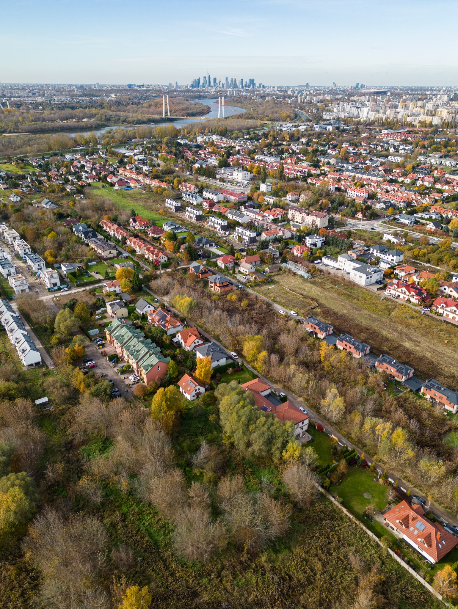 Panoramiczny widok z lotu ptaka na osiedle domów jednorodzinnych z czerwonymi dachami, w tle rzeka z mostem i panorama miasta z wieżowcami, jesienna kolorystyka drzew.