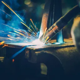 Intensywne spawanie metalu z iskrami i dymem, uchwycone w dynamicznym ujęciu z bliska, widoczna dysza spawarki i fragment rękawicy ochronnej.