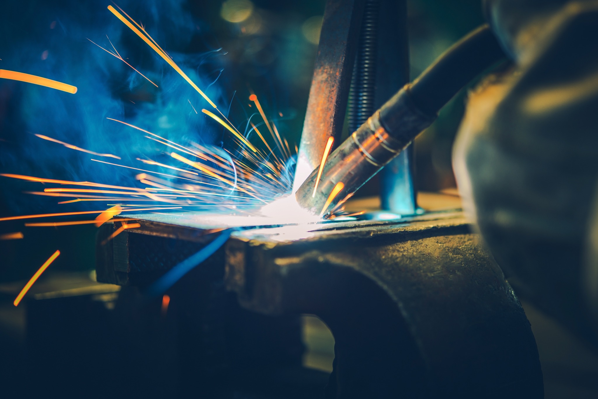 Intensywne spawanie metalu z iskrami i dymem, uchwycone w dynamicznym ujęciu z bliska, widoczna dysza spawarki i fragment rękawicy ochronnej.
