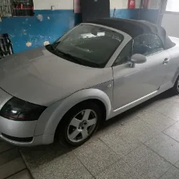 Srebrny Audi TT Roadster z czarnym dachem zaparkowany w warsztacie, widoczne narzędzia na ścianie w tle.