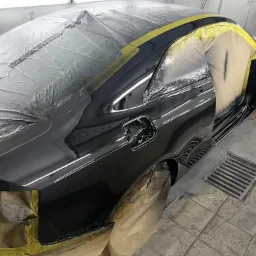 Błyszczący, czarny samochód częściowo zakryty folią ochronną i taśmą malarską podczas lakierowania w warsztacie, widoczna kratka ściekowa w podłodze.