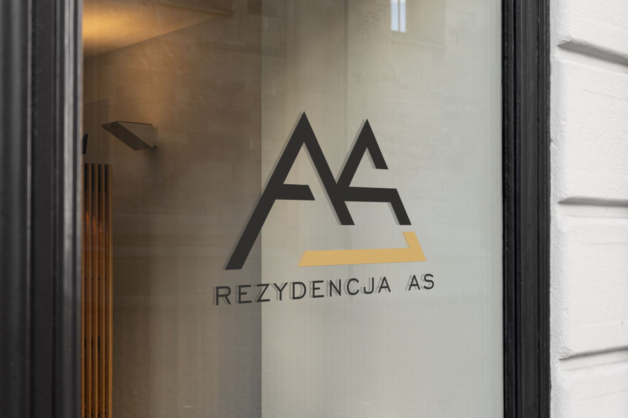 Logo 'Rezydecja AS' umieszczone na szklanych drzwiach wejściowych, widok z zewnątrz, czarna rama drzwi, biała ściana budynku z fakturą.