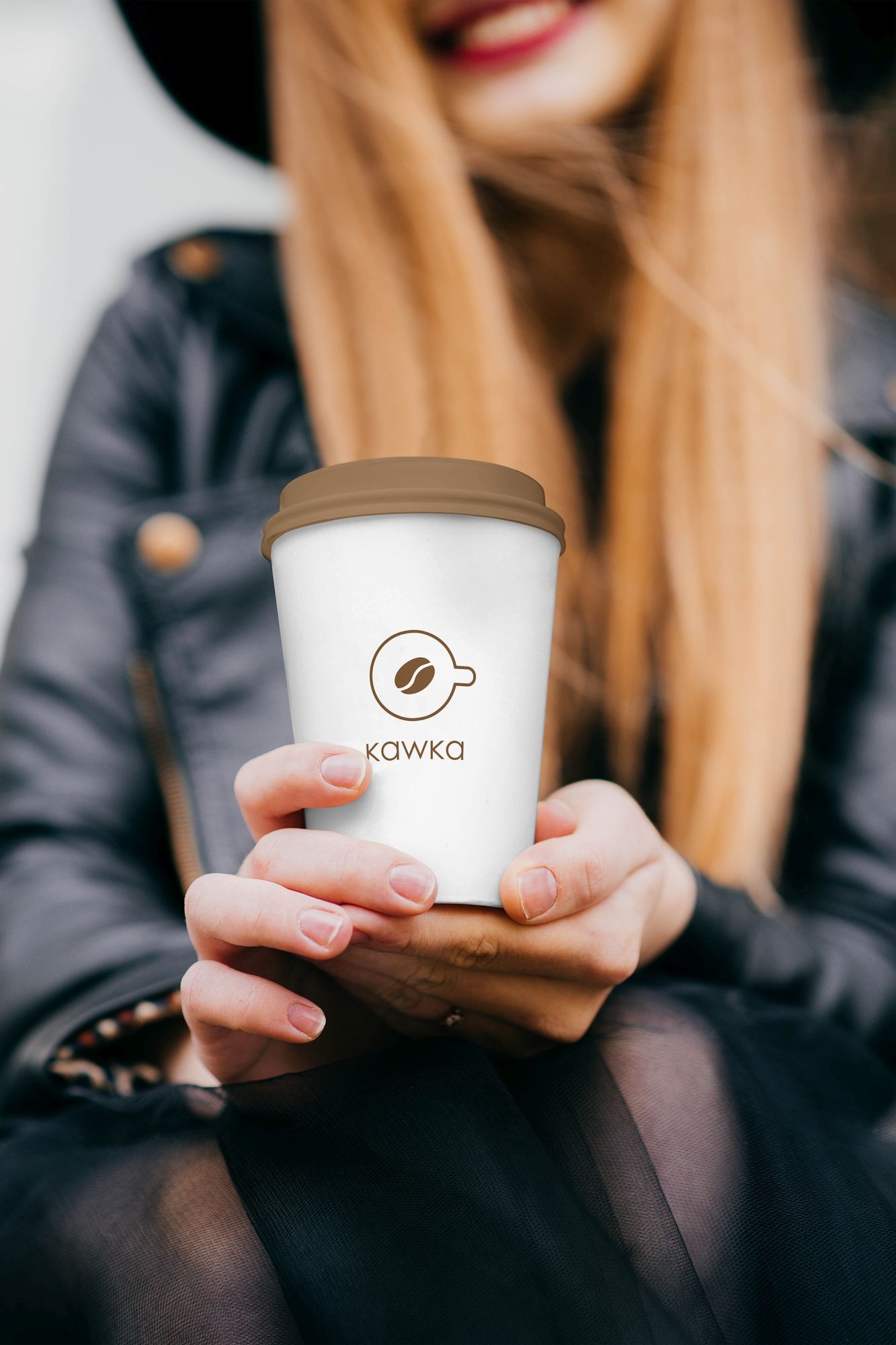 Zbliżenie na dłonie kobiety trzymającej biały kubek z brązową pokrywką, na którym widnieje brązowe logo z ziarnem kawy i napisem 'kawka', w tle rozmyta postać kobiety w czarnym kapeluszu...