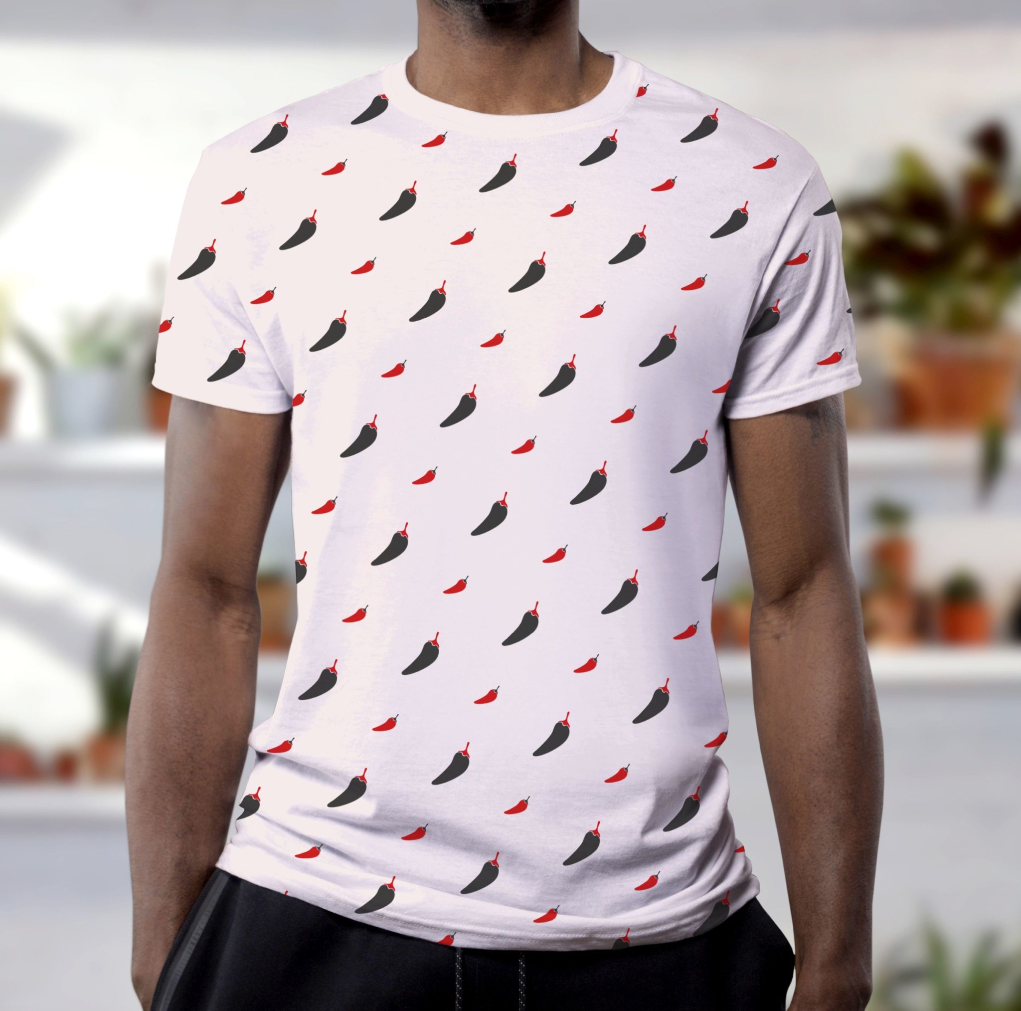 Mężczyzna w białej koszulce z powtarzającym się wzorem czarnych i czerwonych papryczek chili, na rozmytym tle regału z roślinami doniczkowymi.