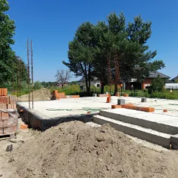 Wylany fundament domu w trakcie budowy, widoczne zbrojenia, cegły, izolacja przeciwwilgociowa, hałda ziemi i fragmenty przyszłych ścian.