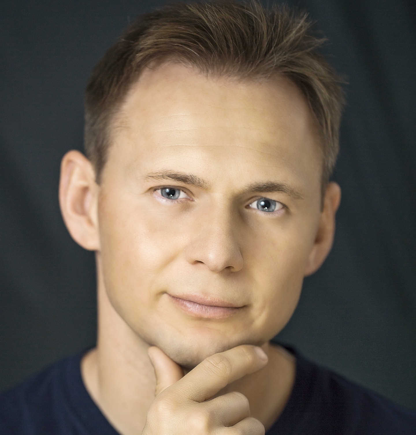 Portret młodego mężczyzny z jasnymi włosami i niebieskimi oczami, zamyślony wyraz twarzy, podpierającego brodę palcem na ciemnym tle.