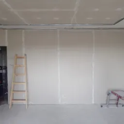 Ściana z płyt kartonowo-gipsowych w trakcie montażu, widoczne profile metalowe na suficie, drewniana drabina oparta o ścianę, pomieszczenie w stanie surowym.