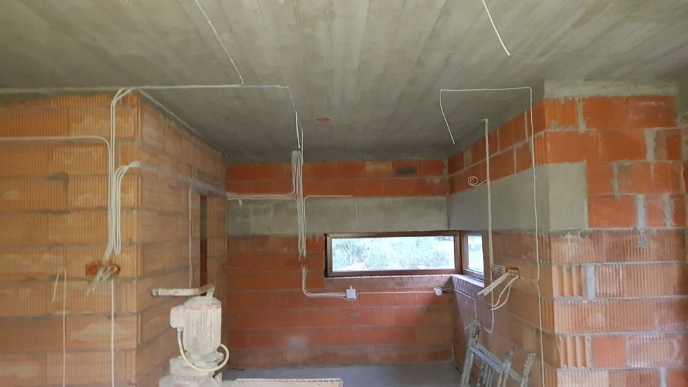 Surowy stan wnętrza budynku w trakcie prac instalacyjnych, widoczne kable elektryczne prowadzone po ścianach z cegły i pod sufitem betonowym.